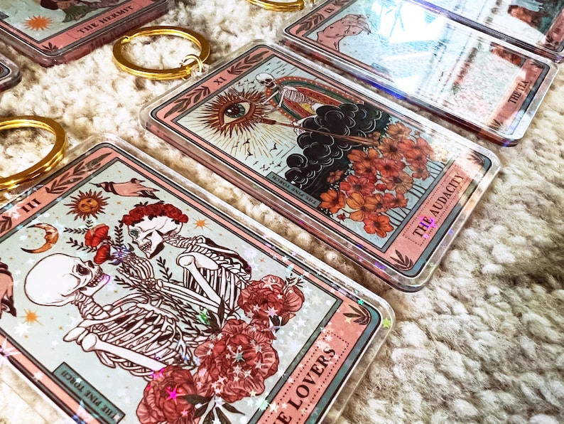 May include: A set of six clear acrylic keychains featuring tarot card designs. Each keychain has a pink border and a different tarot card image, including The Lovers, The Audacity, and The Hermit. The keychains have a glittery effect.