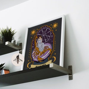 May include: A framed print of a tarot card depicting the Scorpio zodiac sign. The card features a woman sitting on a scorpion with a purple and gold background. The text "Scorpio" is written at the bottom of the card.
