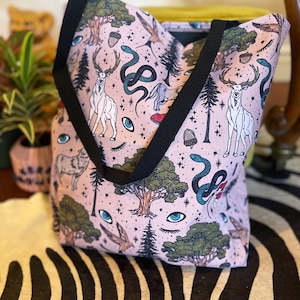 May include: A pink tote bag with a black strap features a whimsical pattern of trees, deer, snakes, and eyes. The pattern is repeated throughout the bag.