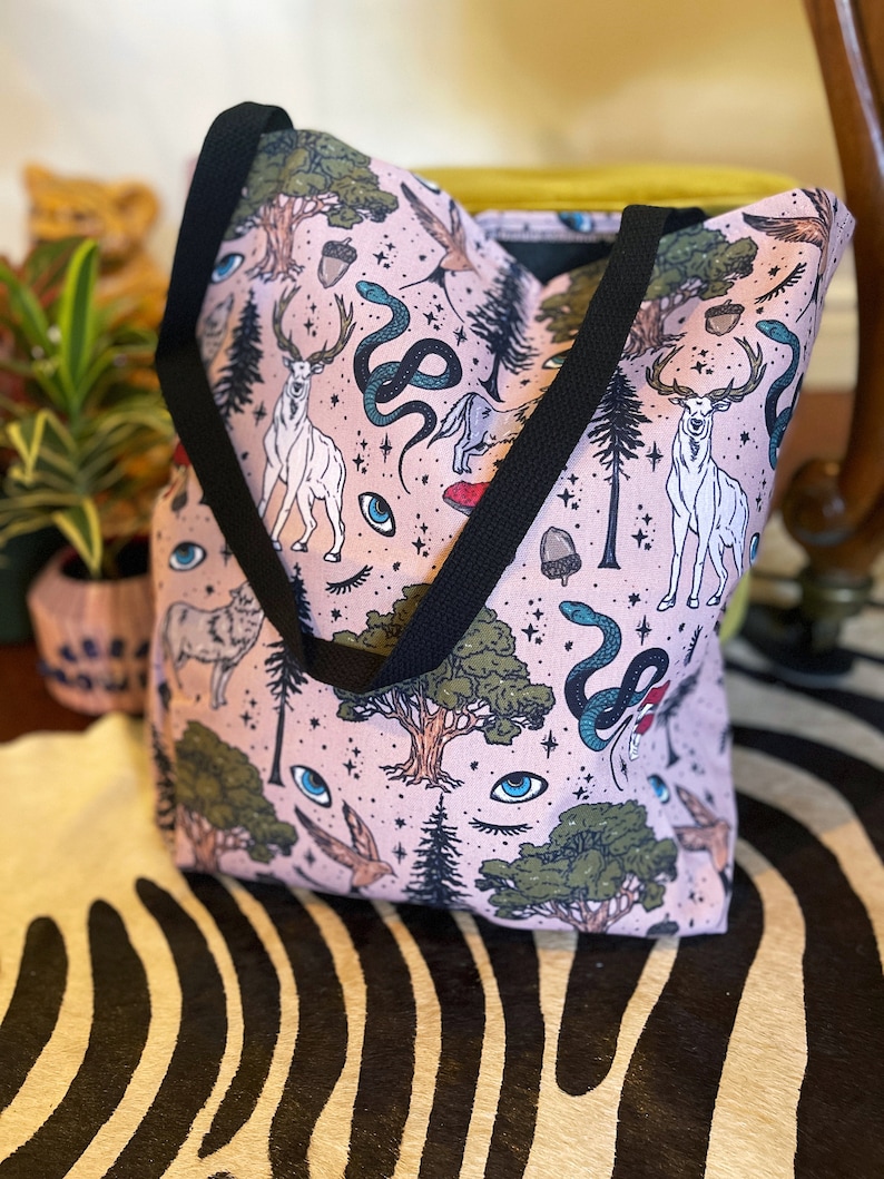 May include: A pink tote bag with a black strap features a whimsical pattern of trees, deer, snakes, and eyes. The pattern is repeated throughout the bag.