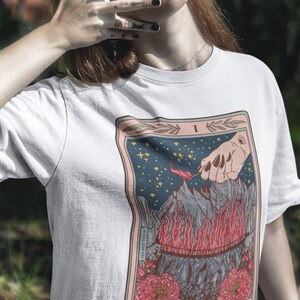 May include: White t-shirt with a tarot card design. The card is titled "The Burned Bridge" and depicts a hand holding a burning torch over a bridge engulfed in flames. The card is surrounded by a pink border with floral accents.