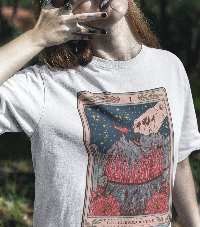 May include: White t-shirt with a tarot card design. The card is titled "The Burned Bridge" and depicts a hand holding a burning torch over a bridge engulfed in flames. The card is surrounded by a pink border with floral accents.