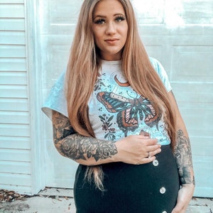 May include: A pregnant person wearing a white t-shirt with a butterfly and moon graphic. The t-shirt has the text "Grow" on the sleeve. The person is also wearing a black skirt with buttons down the front.