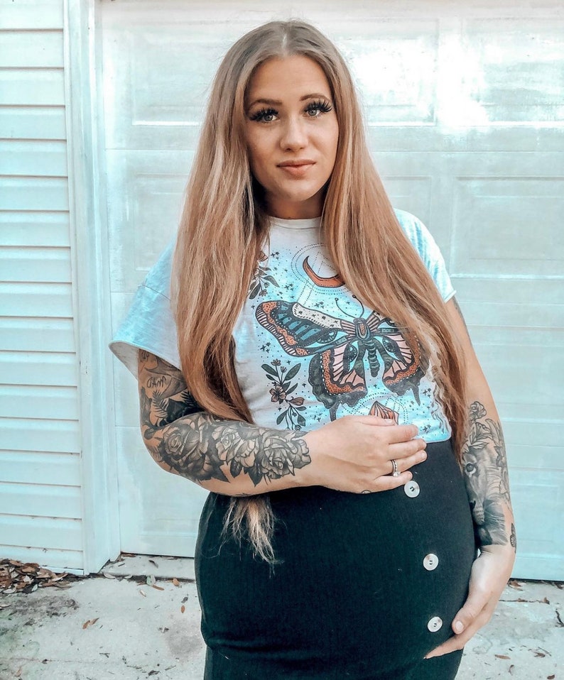 May include: A pregnant person wearing a white t-shirt with a butterfly and moon graphic. The t-shirt has the text "Grow" on the sleeve. The person is also wearing a black skirt with buttons down the front.