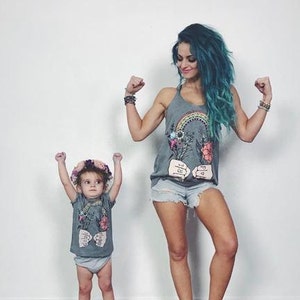 May include: Two people wearing gray tank tops with a rainbow, flowers, and the text "Rise and Shine" printed on them. The person on the left is a child wearing a flower crown. Both people are flexing their biceps.