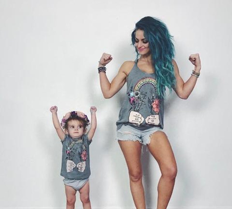 May include: Two people wearing gray tank tops with a rainbow, flowers, and the text "Rise and Shine" printed on them. The person on the left is a child wearing a flower crown. Both people are flexing their biceps.