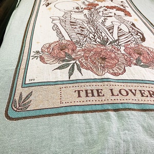 May include: A woven throw blanket featuring a design with two skeletons, flowers, and celestial elements. The words "THE LOVER" are displayed on the blanket. The blanket has a light green background with a fringed edge.