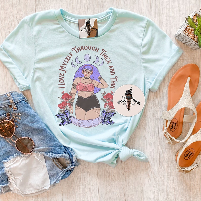 Puede incluir: Camiseta azul claro con un gr&aacute;fico de una mujer con cabello morado y un mensaje positivo sobre el cuerpo que dice "Me amo a m&iacute; misma en la gordura y en la delgadez".