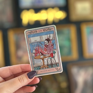 May include: A tarot card with a blue and pink background. The card is numbered XIX and titled "The Zoom Meeting". The card depicts a skeleton sitting at a table with a laptop and a cup of coffee. The skeleton is surrounded by swords that are on fire.