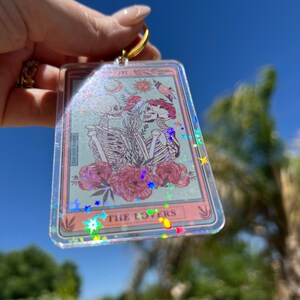 May include: A clear keychain with a pink and blue tarot card design. The card features two skeletons holding hands with flowers and the text "THE LOVERS". The keychain is covered in iridescent glitter.