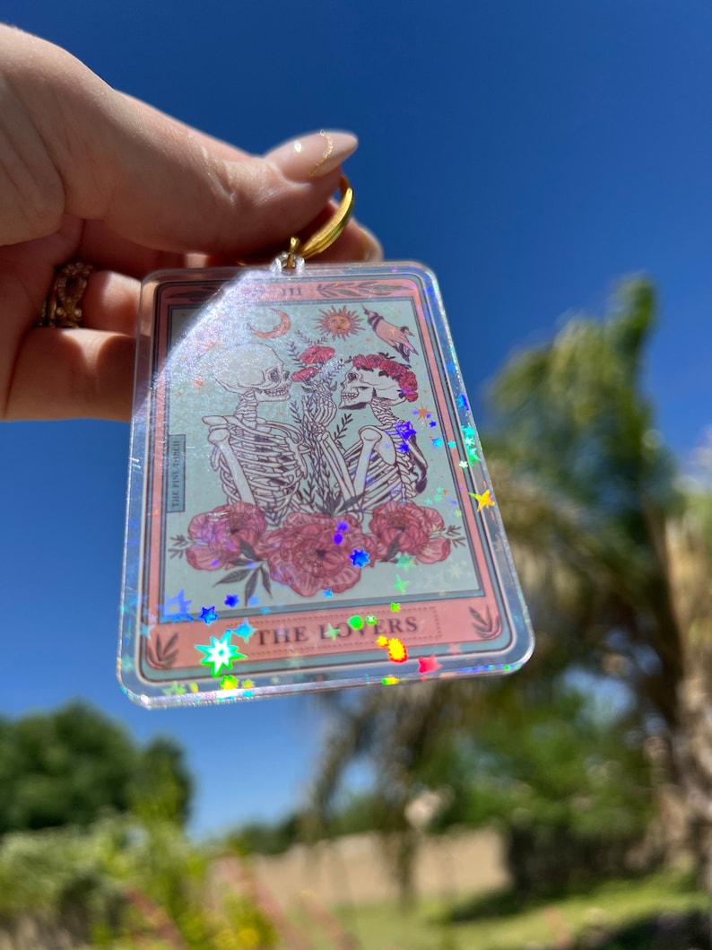 May include: A clear keychain with a pink and blue tarot card design. The card features two skeletons holding hands with flowers and the text "THE LOVERS". The keychain is covered in iridescent glitter.