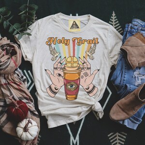 Op de afbeelding: Een beige T-shirt met de tekst "Holy Grail" bedrukt in oranje en bruin. De afbeelding toont een koffiebeker met een oog en twee handen die hem omhoog houden.