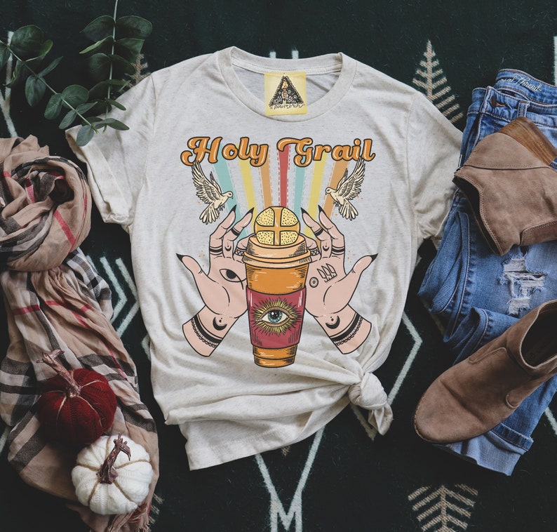 Op de afbeelding: Een beige T-shirt met de tekst "Holy Grail" bedrukt in oranje en bruin. De afbeelding toont een koffiebeker met een oog en twee handen die hem omhoog houden.