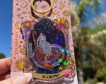 May include: A holographic keychain featuring the zodiac sign Scorpio. The keychain is a clear plastic charm with a gold-colored border and a black and white illustration of a woman sitting on a scorpion. The background is a dark blue with stars and glitter. The text "Scorpio" is printed in gold on the charm.
