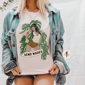 K&ouml;nnte beinhalten: Ein pinkfarbenes T-Shirt mit einem Aufdruck einer Frau in einer Dschungelkulisse. Die Frau tr&auml;gt einen Leopardenrock und ein wei&szlig;es Oberteil. Der Text "SEND NODES" ist auf dem Shirt aufgedruckt.
