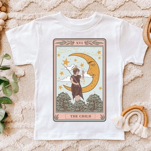 May include: White t-shirt with a vintage tarot card design featuring a child standing on a crescent moon with stars in the background. The card reads "XVI THE CHILD".