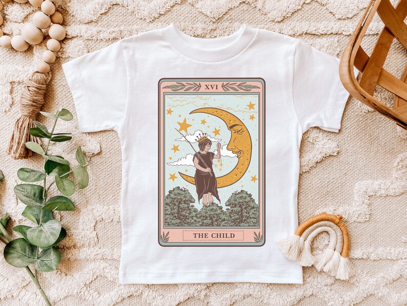 May include: White t-shirt with a vintage tarot card design featuring a child standing on a crescent moon with stars in the background. The card reads "XVI THE CHILD".