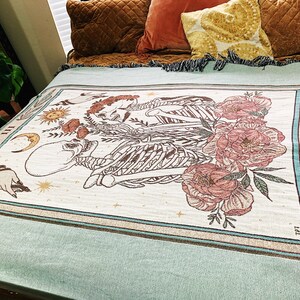 May include: Cream-colored woven throw blanket featuring a tarot card design with skeletons, flowers, and celestial elements. The border is teal with the words "THE LOVERS". The blanket is draped over a bed with pillows.