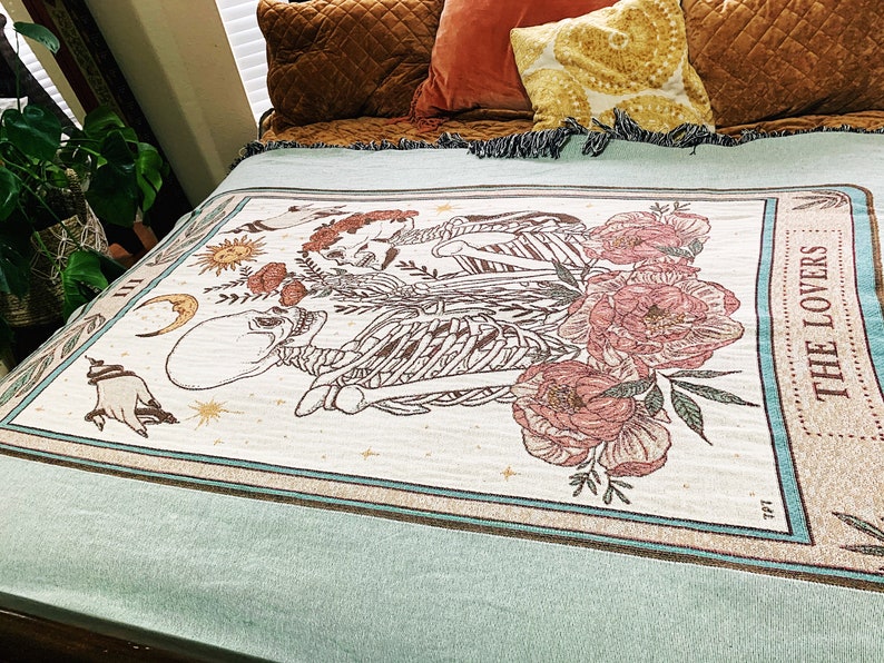 May include: Cream-colored woven throw blanket featuring a tarot card design with skeletons, flowers, and celestial elements. The border is teal with the words "THE LOVERS". The blanket is draped over a bed with pillows.