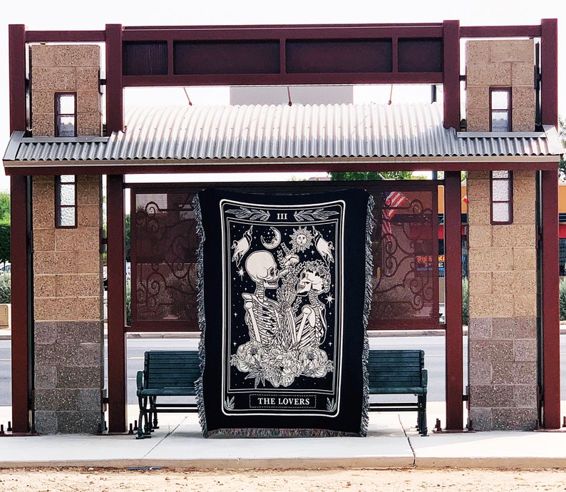 May include: Black and white tapestry featuring a skeletal couple with floral accents, a moon, and a sun. The tapestry is labeled "THE LOVERS" and is displayed outdoors. The design is framed by a decorative border.
