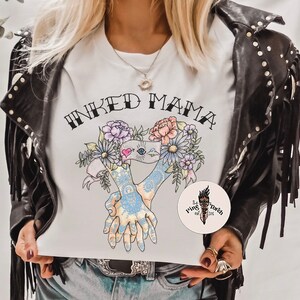 Pu&ograve; includere: T-shirt bianca con la scritta "INKED MAMA" sopra un disegno floreale e due mani tatuate che si tengono. &Egrave; visibile un adesivo rotondo con il logo di una torcia.
