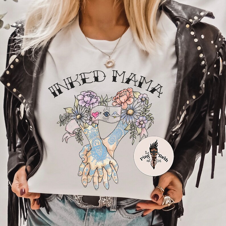 Pu&ograve; includere: T-shirt bianca con la scritta "INKED MAMA" sopra un disegno floreale e due mani tatuate che si tengono. &Egrave; visibile un adesivo rotondo con il logo di una torcia.