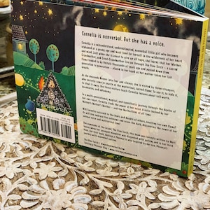 May include: The back cover of a children's book titled "Cornelia and the Fine Torch." The book features a colorful illustration of a girl holding a torch. The text describes the story of a nonverbal girl who finds her voice.