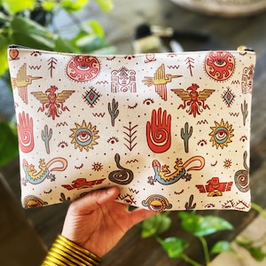May include: Cream-colored zippered pouch with a repeating pattern of Southwestern-inspired illustrations in red, yellow, blue, and black. The design includes suns, birds, hands, and other symbols. The pouch is held in a hand with gold bangles.