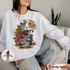 May include: White sweatshirt with a graphic of a skeleton holding a watering can and surrounded by flowers and mushrooms. The text "SELF CARE" is on a banner below the skeleton.