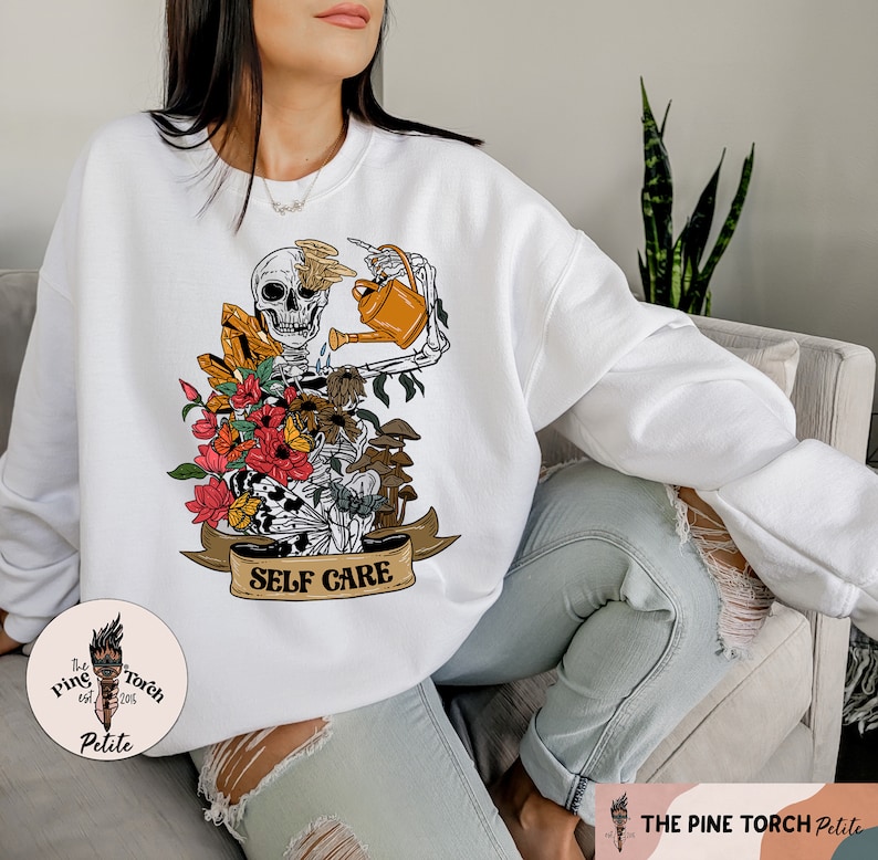 May include: White sweatshirt with a graphic of a skeleton holding a watering can and surrounded by flowers and mushrooms. The text "SELF CARE" is on a banner below the skeleton.