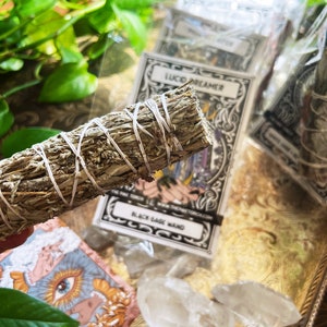 Könnte beinhalten: Ein Bündel getrockneter Salbei, auch bekannt als Räucherstäbchen, mit Schnur gebunden. Der Salbei ist hellgrau und von klaren Quarzkristallen und kleinen Karten mit dem Text "Lucid Dreamer" und "Black Sage Wand" umgeben.