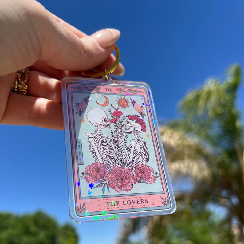May include: A pink and blue tarot card keychain with a holographic finish. The card features a skeletal couple holding flowers and the text "The Lovers" and "III".
