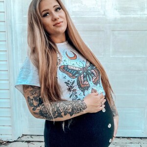 May include: A woman wearing a white t-shirt with a butterfly and moon graphic. The shirt has a floral design and the butterfly has blue, pink, and orange wings. The woman has long blonde hair and is wearing a black skirt with buttons down the front. She has a tattoo on her arm of roses and other flowers.