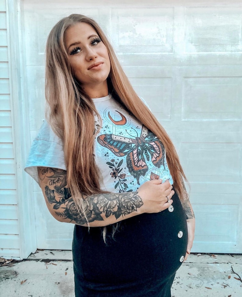 May include: A woman wearing a white t-shirt with a butterfly and moon graphic. The shirt has a floral design and the butterfly has blue, pink, and orange wings. The woman has long blonde hair and is wearing a black skirt with buttons down the front. She has a tattoo on her arm of roses and other flowers.