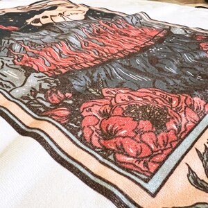 May include: A vintage-style tarot card with a blue border and a peach background. The card features a mountain scene with flames and a skull, and a large red flower in the bottom right corner. The text "THE..." is visible in the bottom right corner.