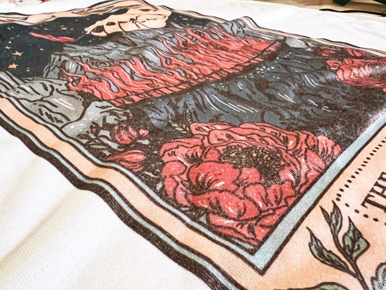 May include: A vintage-style tarot card with a blue border and a peach background. The card features a mountain scene with flames and a skull, and a large red flower in the bottom right corner. The text "THE..." is visible in the bottom right corner.