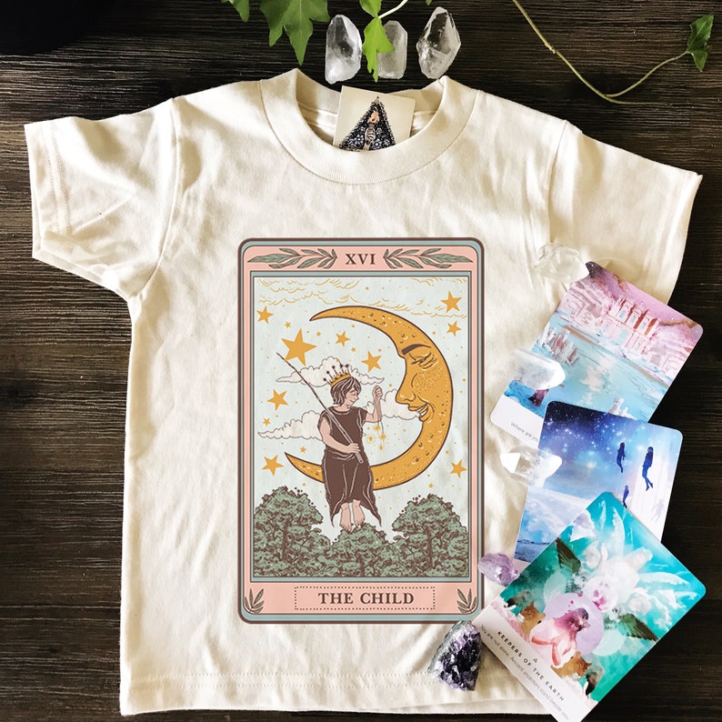 May include: A white t-shirt with a vintage tarot card design featuring The Child card. The card depicts a person standing on a crescent moon with stars in the background. The text "XVI THE CHILD" is printed on the card.