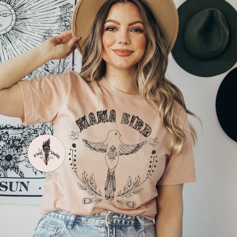 May include: A peach-colored t-shirt with a black graphic of a bird with the text "Mama Bird" above it. The bird has a heart design on its chest and is surrounded by floral designs.
