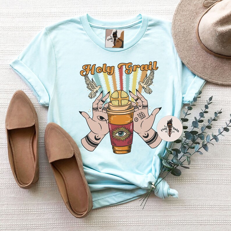 Op de afbeelding: Lichtblauw T-shirt met een afbeelding van een koffiebeker met een oog en handen die deze vasthouden. De tekst "Holy Grail" staat boven de afbeelding.