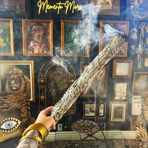 May include: A person holds a white sage smudge stick, burning with smoke, in front of a wall decorated with framed artwork. The wall features a variety of paintings and prints, including a lion, a religious figure, and abstract art. The text "Memento Mori" is visible in yellow neon letters at the top of the wall.