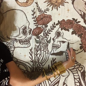 May include: A cream-colored tapestry featuring a skeletal couple with floral accents. The design includes a crescent moon, a sun, and a floral arrangement. A person's arm with gold bangles and a tattoo is visible.