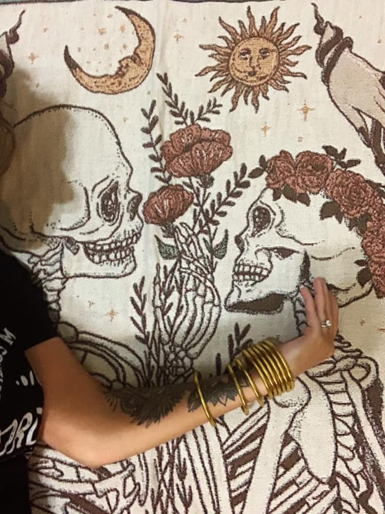 May include: A cream-colored tapestry featuring a skeletal couple with floral accents. The design includes a crescent moon, a sun, and a floral arrangement. A person's arm with gold bangles and a tattoo is visible.