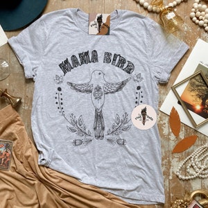 May include: Gray t-shirt with a black and white illustration of a bird with the text "Mama Bird" above it. The bird is surrounded by floral designs.