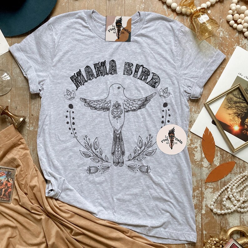 May include: Gray t-shirt with a black and white illustration of a bird with the text "Mama Bird" above it. The bird is surrounded by floral designs.