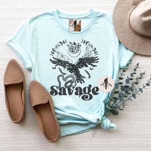 May include: A light blue t-shirt with a black and white graphic design of a raven, snake, and moon phases. The text "Savage" is printed in black below the graphic.