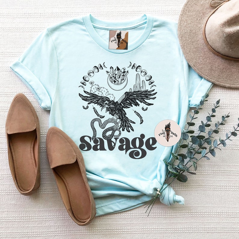 May include: A light blue t-shirt with a black and white graphic design of a raven, snake, and moon phases. The text "Savage" is printed in black below the graphic.