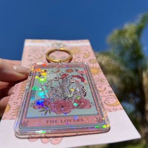May include: A clear keychain with a holographic finish featuring a tarot card design. The card depicts two skeletons holding hands in front of a floral background. The card reads "THE LOVERS".