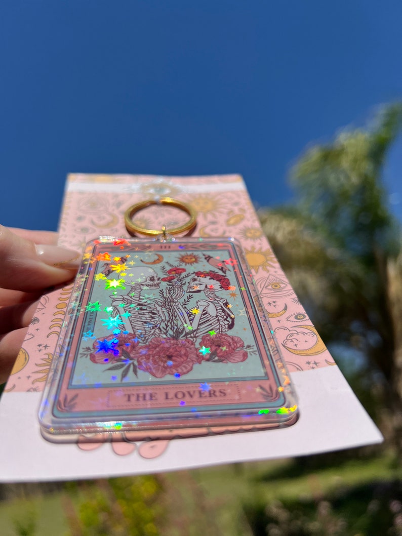 May include: A clear keychain with a holographic finish featuring a tarot card design. The card depicts two skeletons holding hands in front of a floral background. The card reads "THE LOVERS".