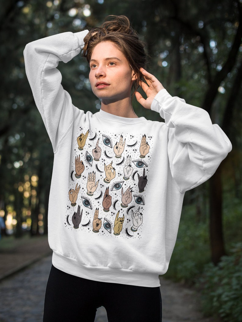 May include: White sweatshirt with a colorful pattern of hands, eyes, and crescent moons. The pattern is repeated throughout the sweatshirt.
