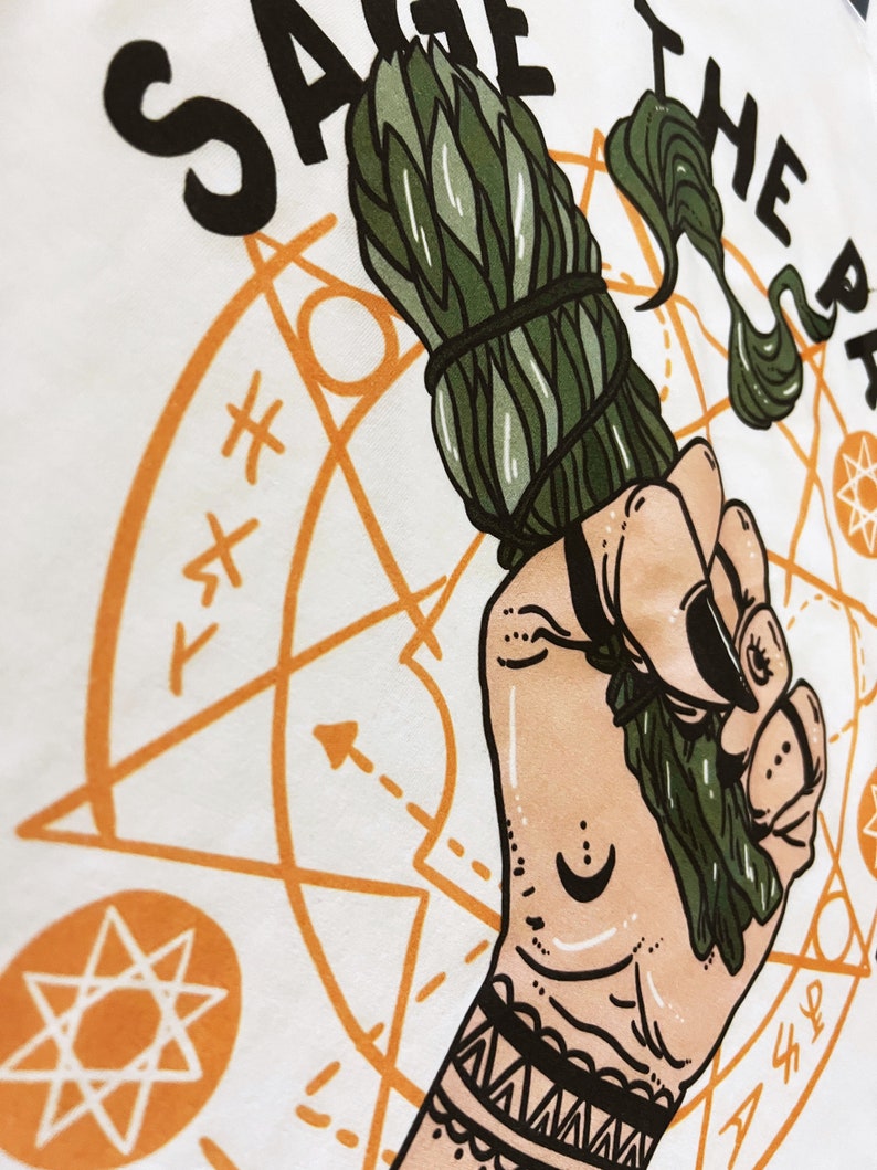 Puede incluir: Una camiseta blanca con una mano sosteniendo un palo de salvia. La mano tiene un tatuaje negro en la mu&ntilde;eca y un tatuaje de luna creciente en la palma. El texto "Sage the Path" est&aacute; impreso en la camiseta en negro.